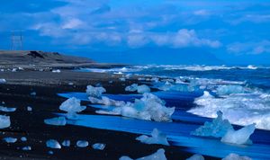 Large chunks of ice from an iceberg are scattered across a black sandy beach with breaking waves. There are large powerlines and mountains in the background.